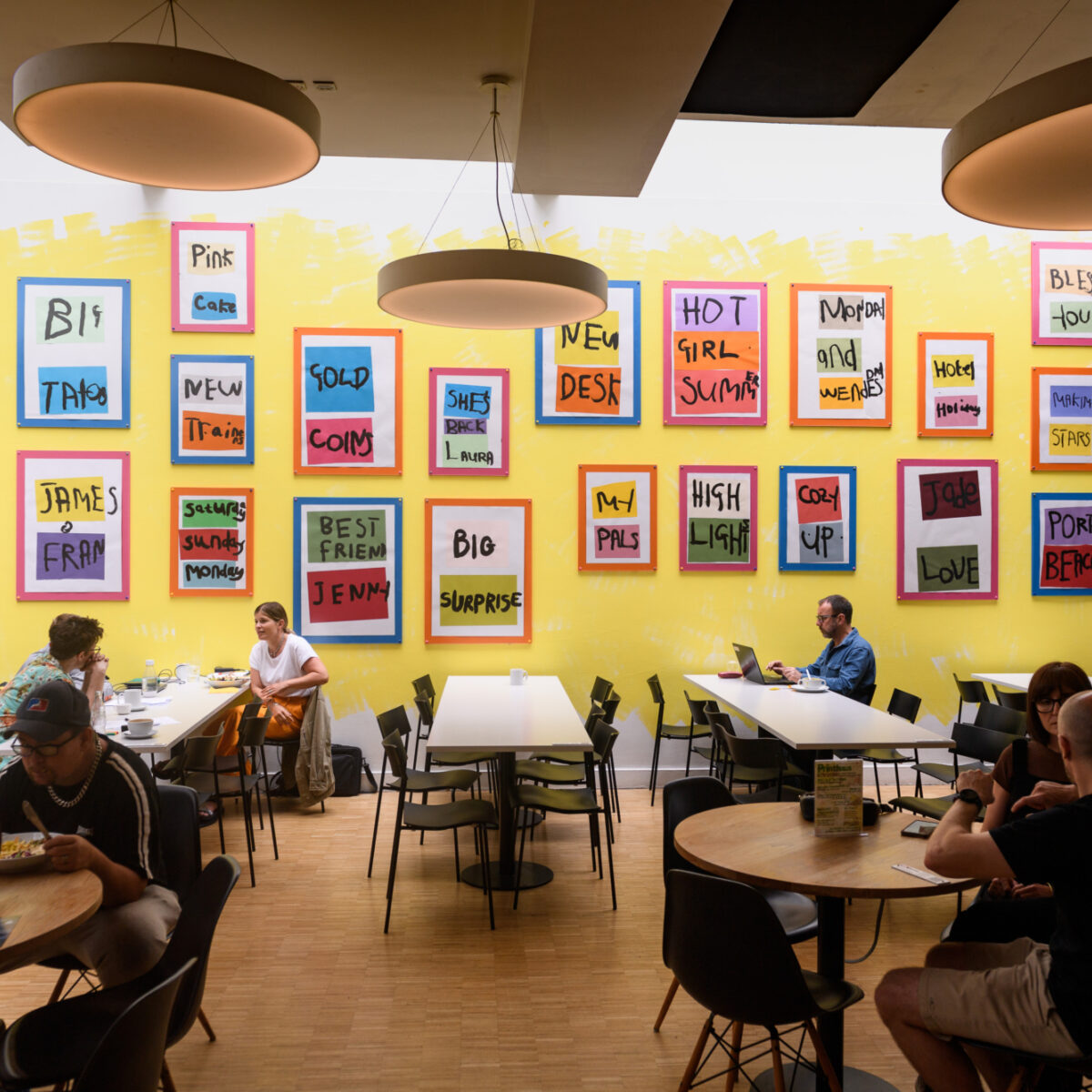 Bright yellow cafe wall in Chapter Arts Centre. Multiple colourful frames hang on the yellow wall, each reading different painted bold text. The cafe is busy with people sat at each table.