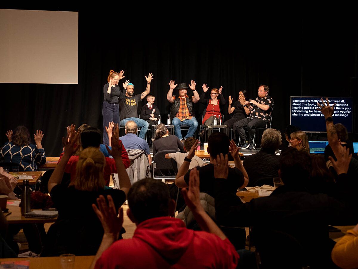 A group of people on stage applaud via BSL, the crowd watching them also applaud via BSL and through clapping.