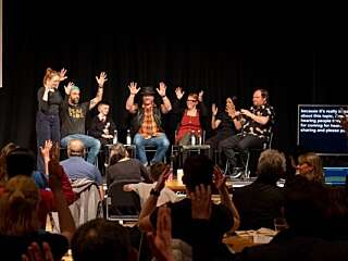 A group of poeple sat on a stage applaud through BSL, as do a crowd of people in front of them.