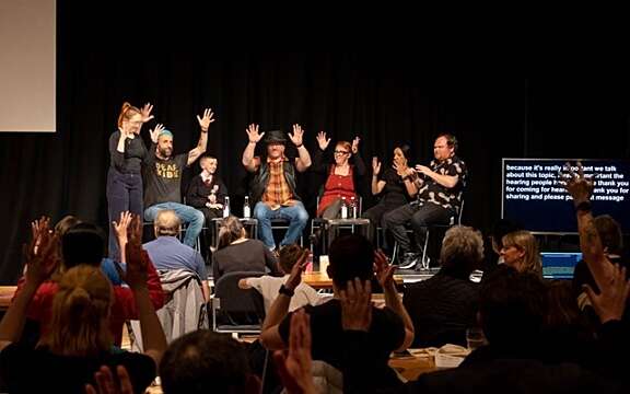 A group of poeple sat on a stage applaud through BSL, as do a crowd of people in front of them.