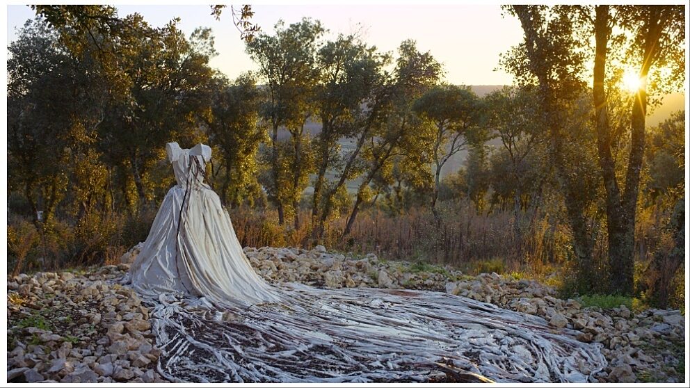 A white dress stands alone on rocky ground in a forest. The sun sets behind the dress.