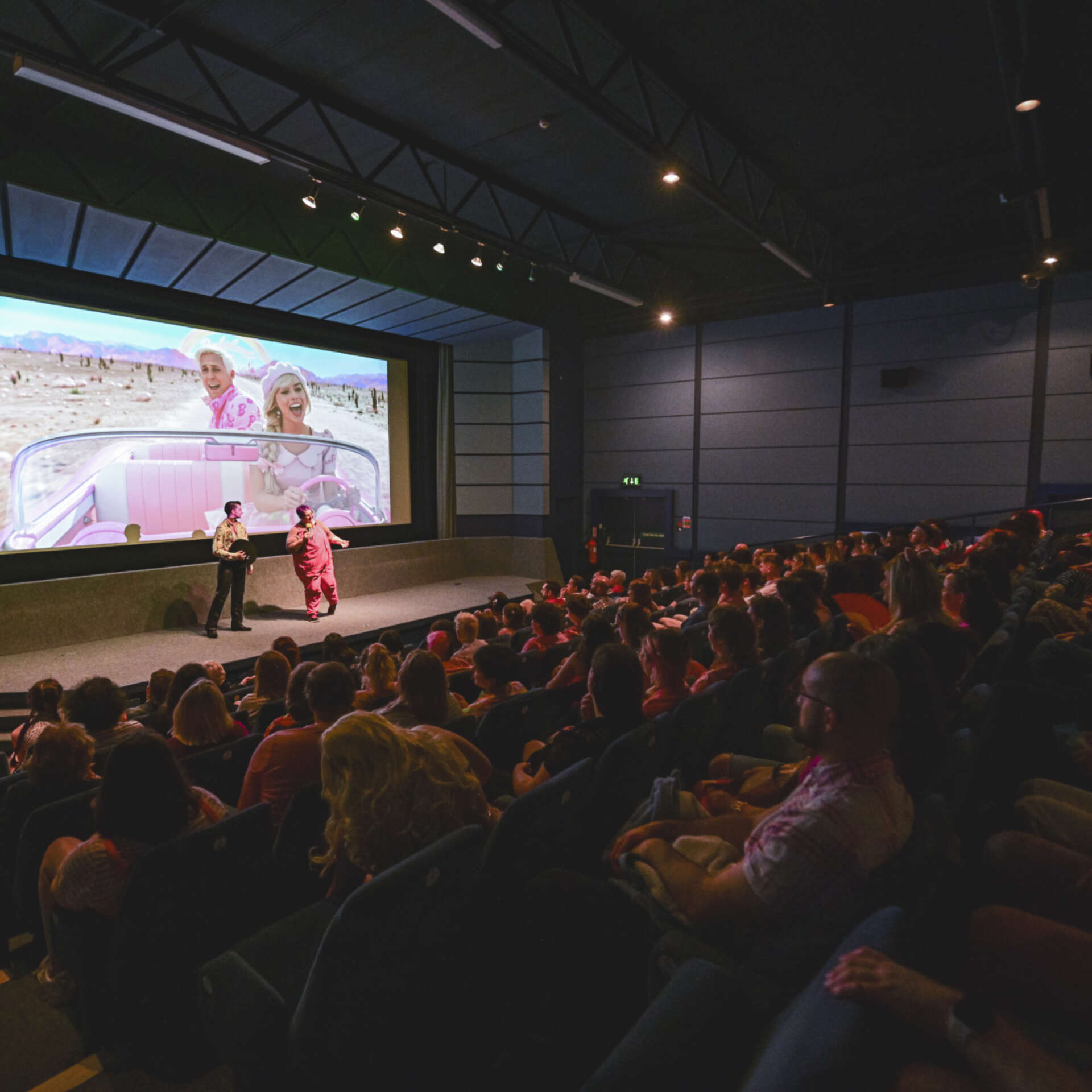 A dimly lit, full cinema. Two people are stood on stage in front of the cinema screen talking to the audience. Behind the speakers is a still image from the Barbie Movie.