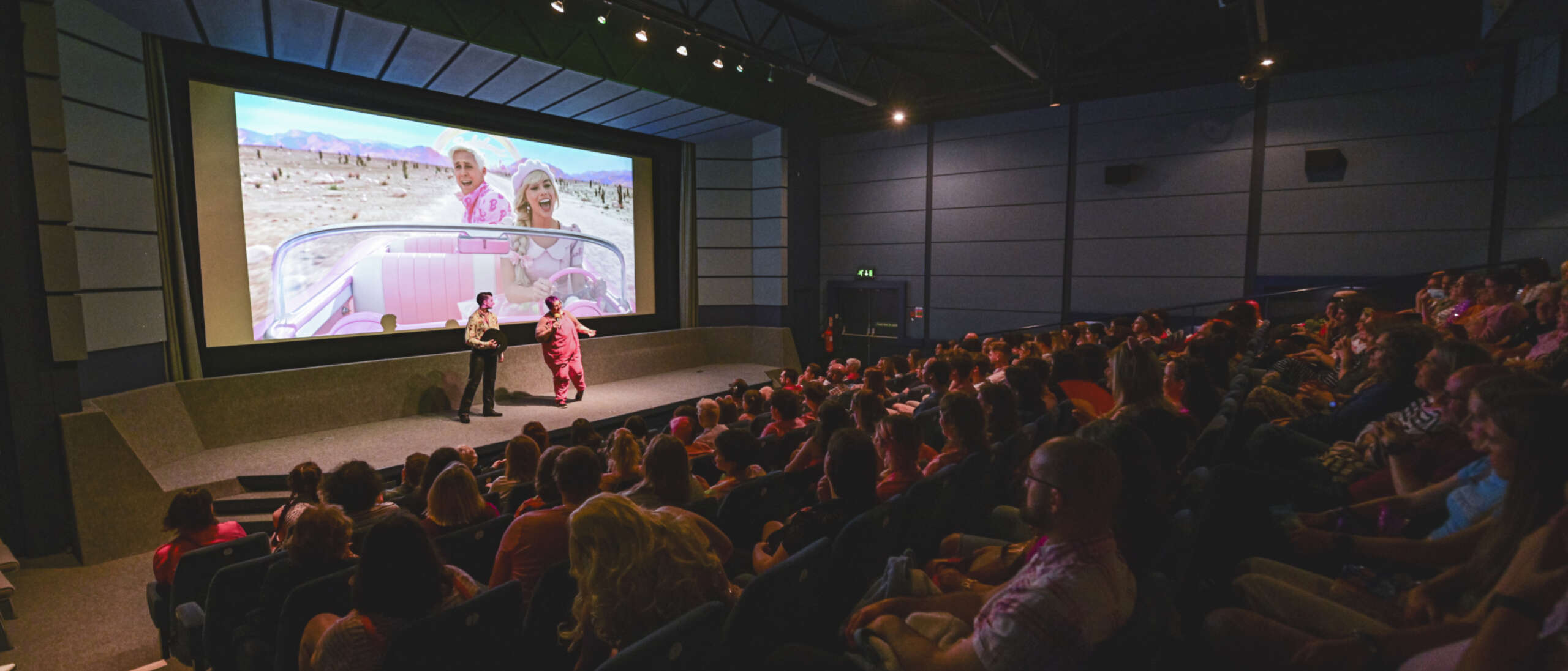 A dimly lit, full cinema. Two people are stood on stage in front of the cinema screen talking to the audience. Behind the speakers is a still image from the Barbie Movie.
