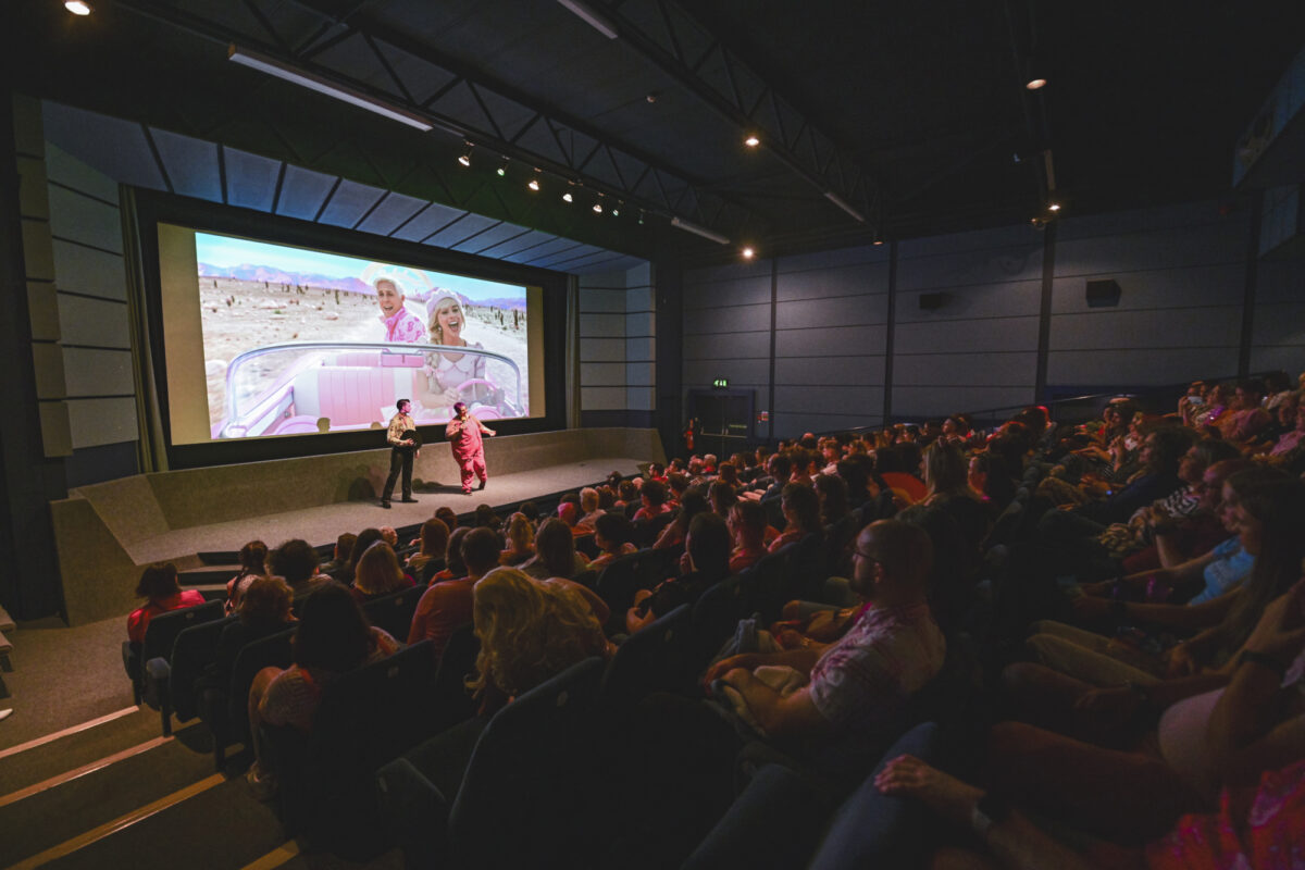 A dimly lit, full cinema. Two people are stood on stage in front of the cinema screen talking to the audience. Behind the speakers is a still image from the Barbie Movie.