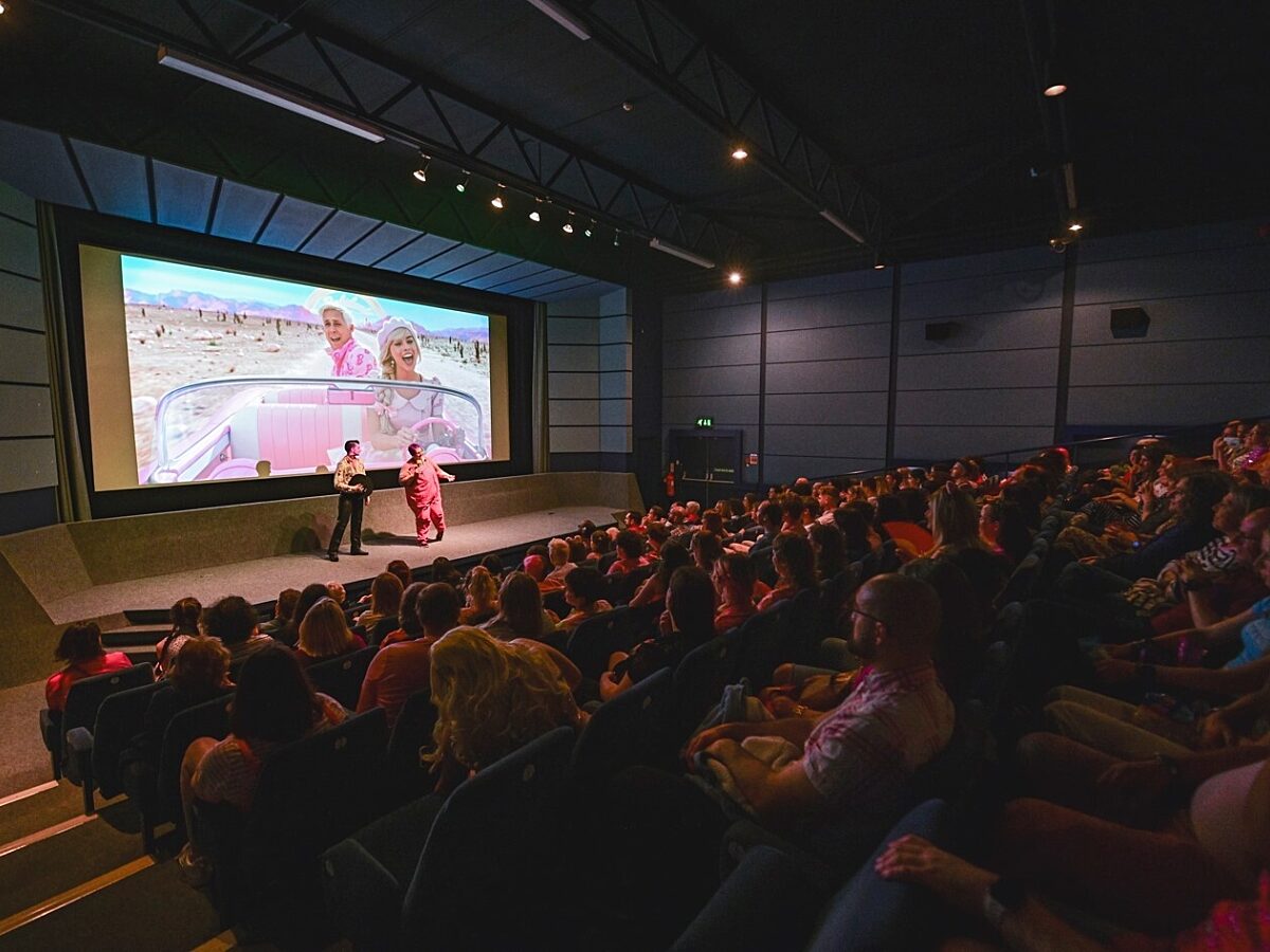 A dimly lit, full cinema. Two people are stood on stage in front of the cinema screen talking to the audience. Behind the speakers is a still image from the Barbie Movie.