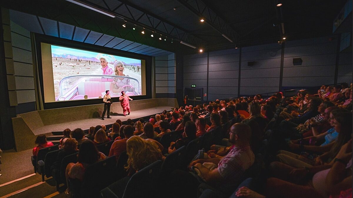 A dimly lit, full cinema. Two people are stood on stage in front of the cinema screen talking to the audience. Behind the speakers is a still image from the Barbie Movie.