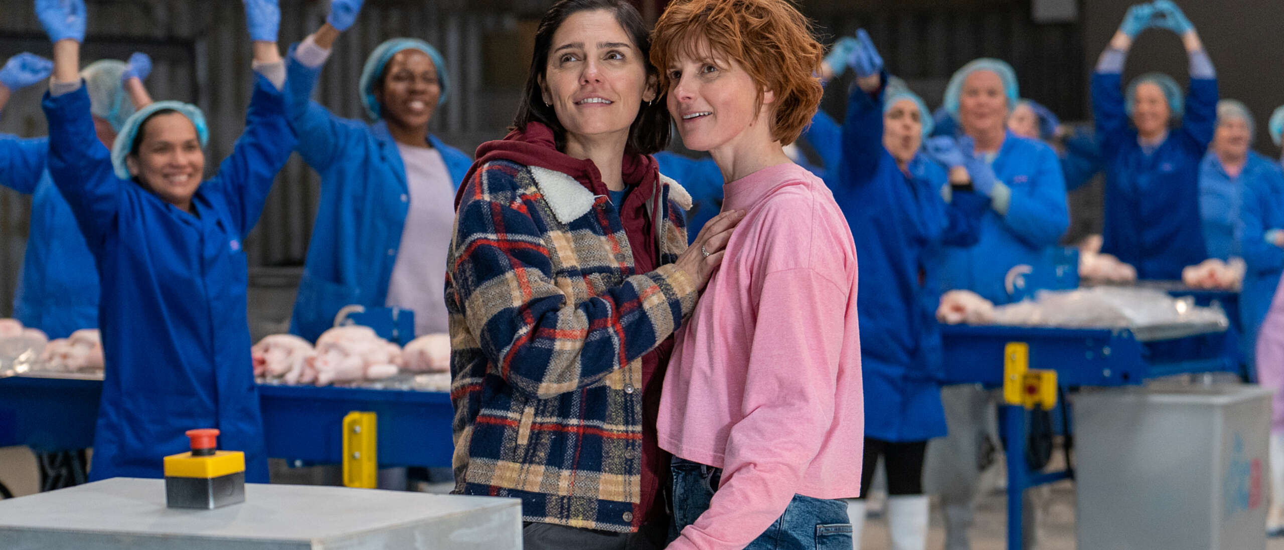 Two women are stood holding each other in a busy factory. They're stood among female factory workers dressed in blue uniform and in front of them is a red button.