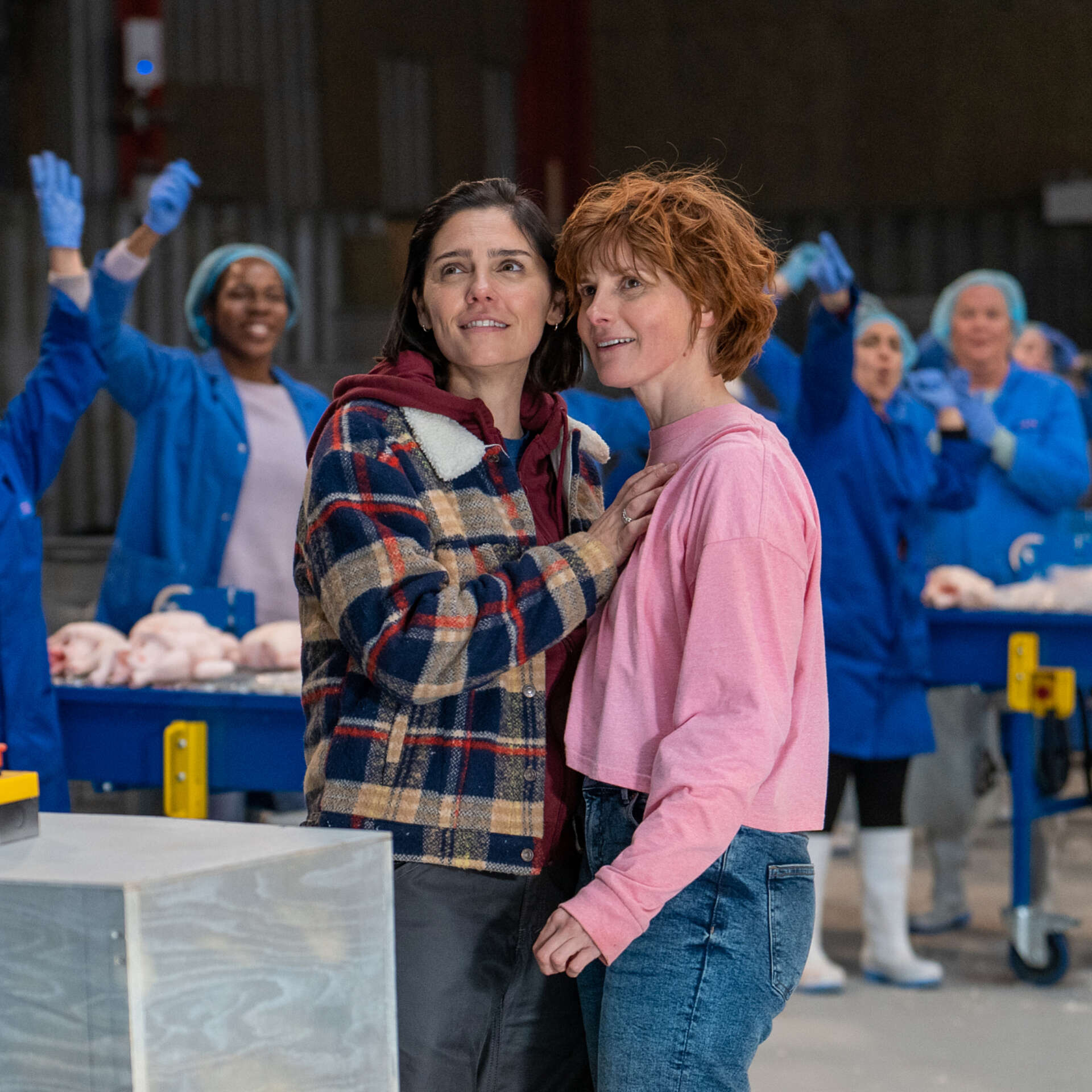Two women are stood holding each other in a busy factory. They're stood among female factory workers dressed in blue uniform and in front of them is a red button.