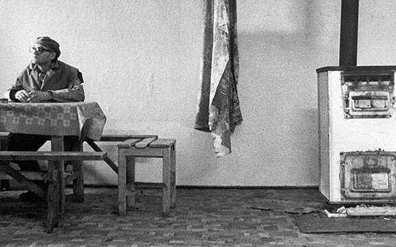 A black and white still image of a man sat at a wooden dining room table in a Hungarian home.