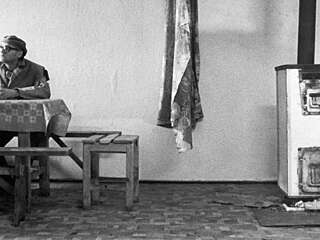 A black and white still image of a man sat at a wooden dining room table in a Hungarian home.