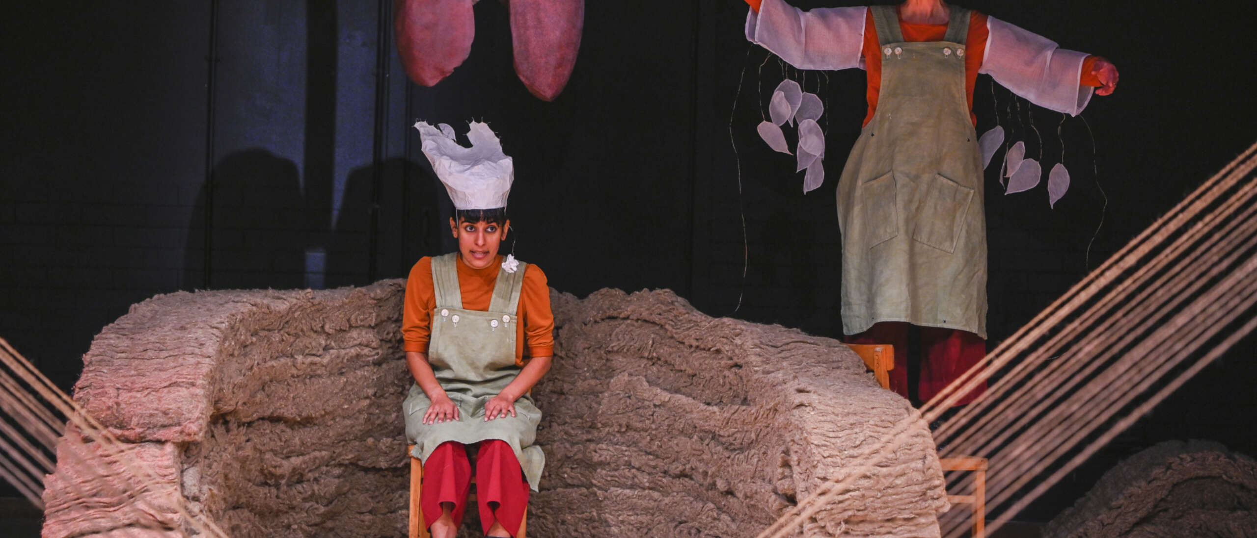 A performer is sat on stage wearing green overalls and a tall white hat. Above the performer is a stage prop of large red lungs. to the right of the performer another person stands on a chair wearing green overalls with tear drops hanging from their arms.