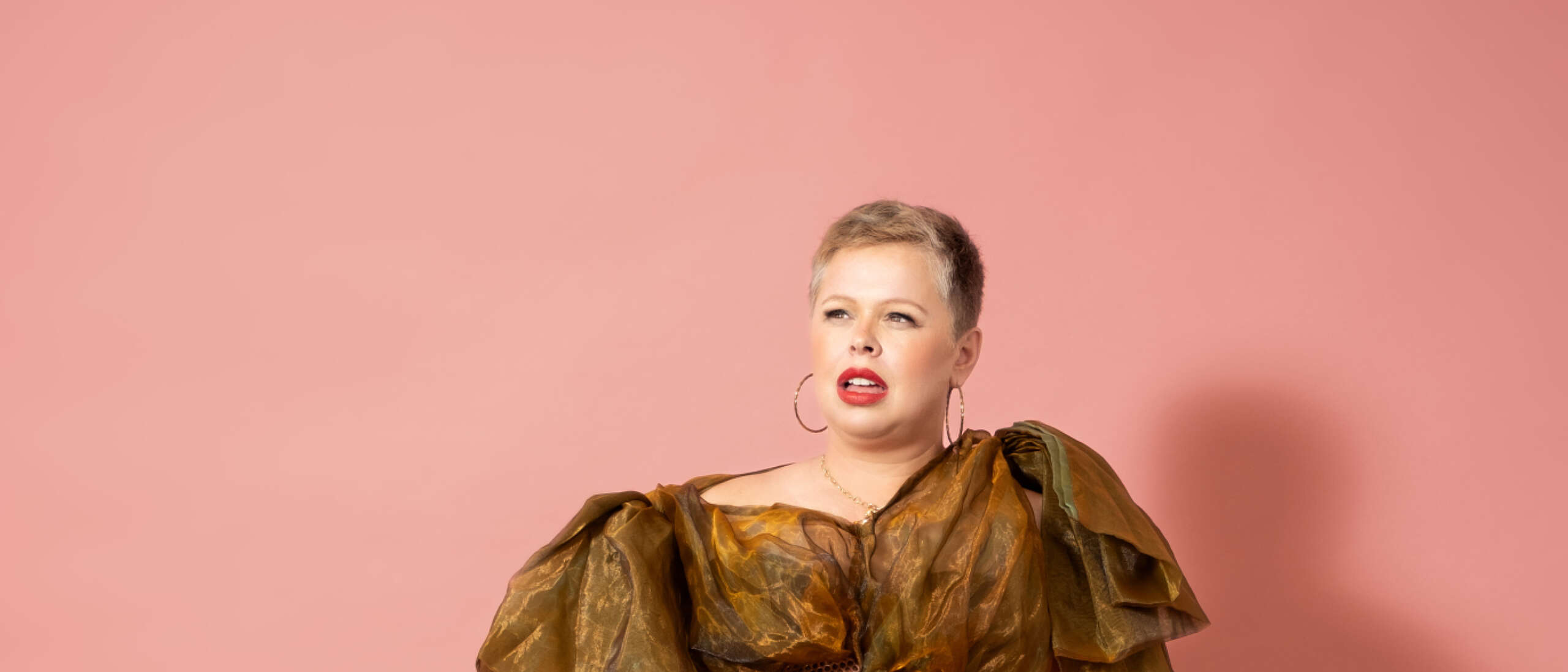 Comedian Laura Smyth Wears a long brown frilled gown and leans on a full washing basket.