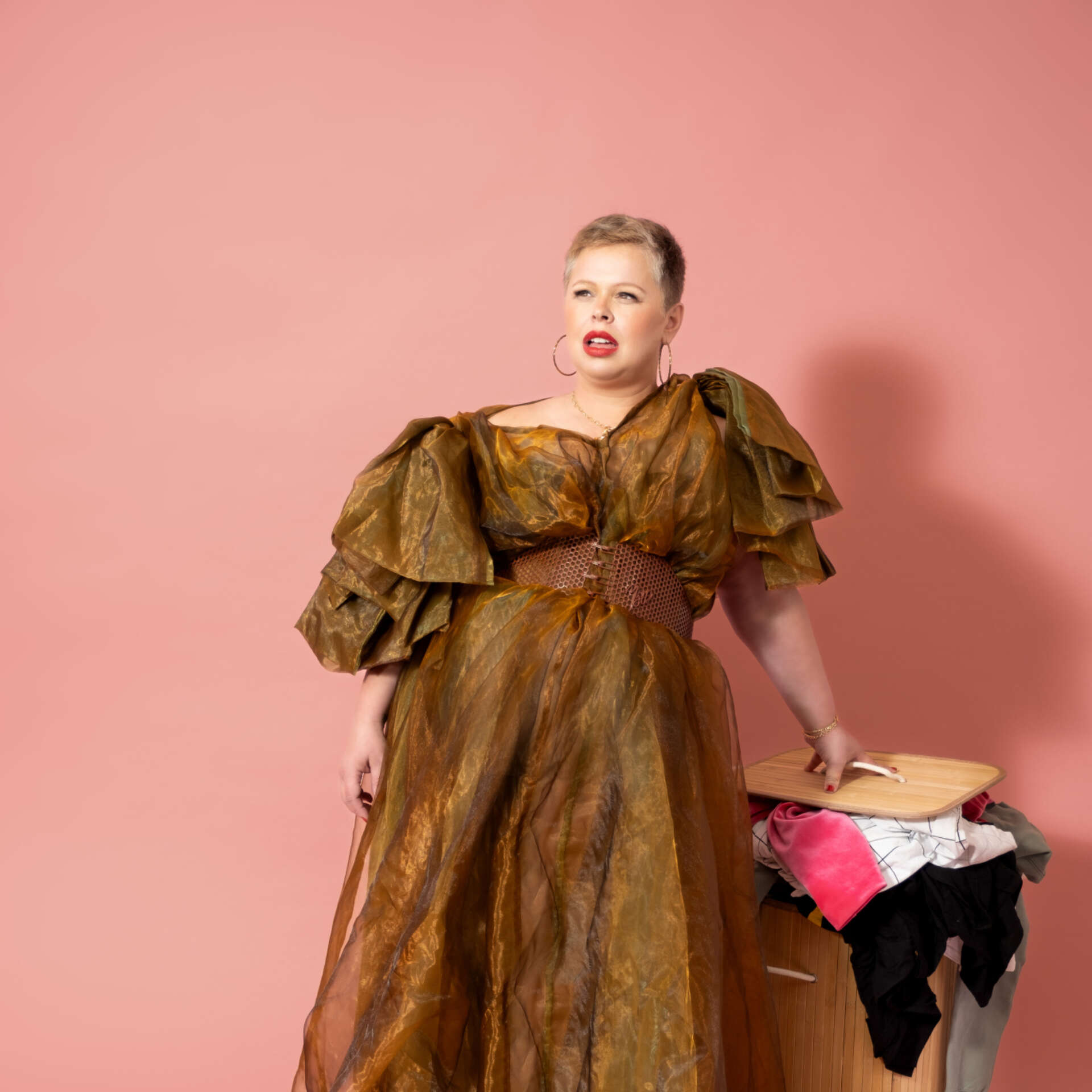 Comedian Laura Smyth Wears a long brown frilled gown and leans on a full washing basket.