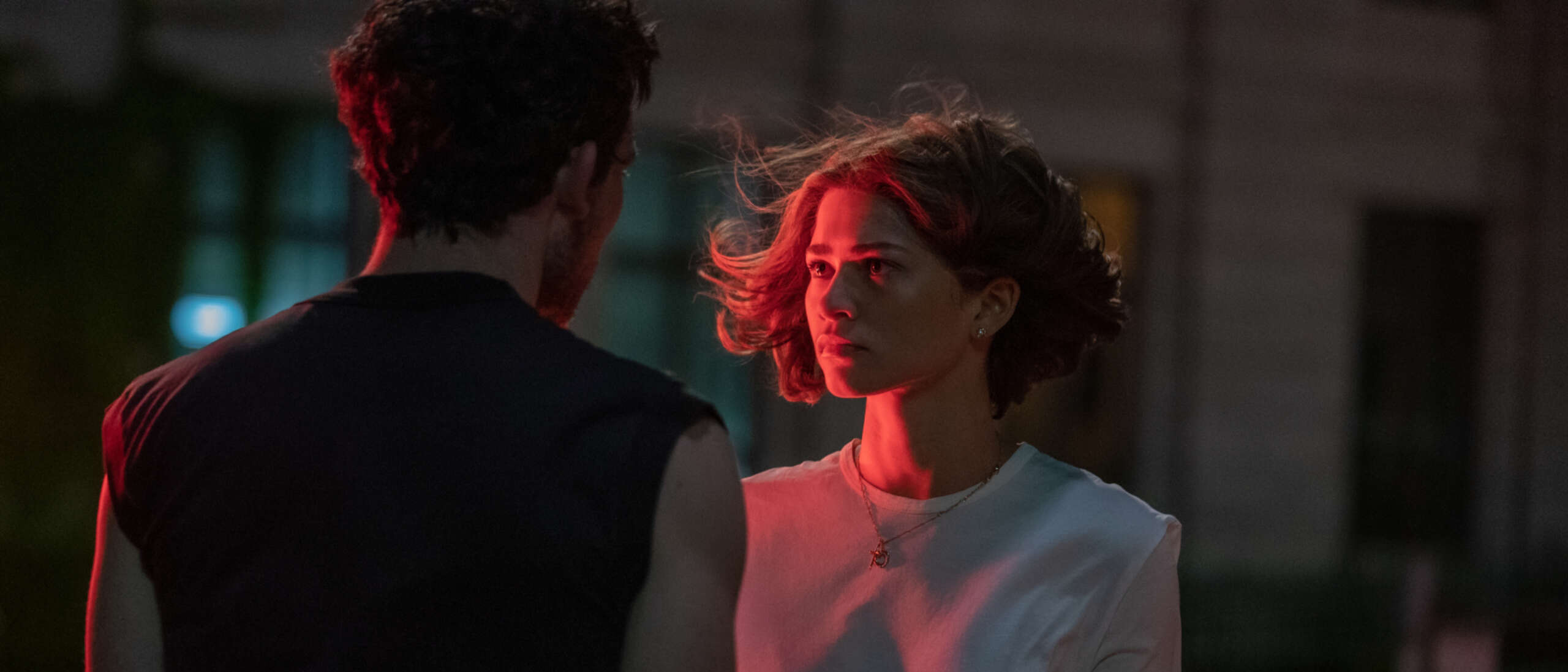 Zendaya and Josh O'Connor star in Challengers. Both are stood facing each other and Josh O'Connor has his back towards the camera. A hot pink light shines on the left side of Zendaya.