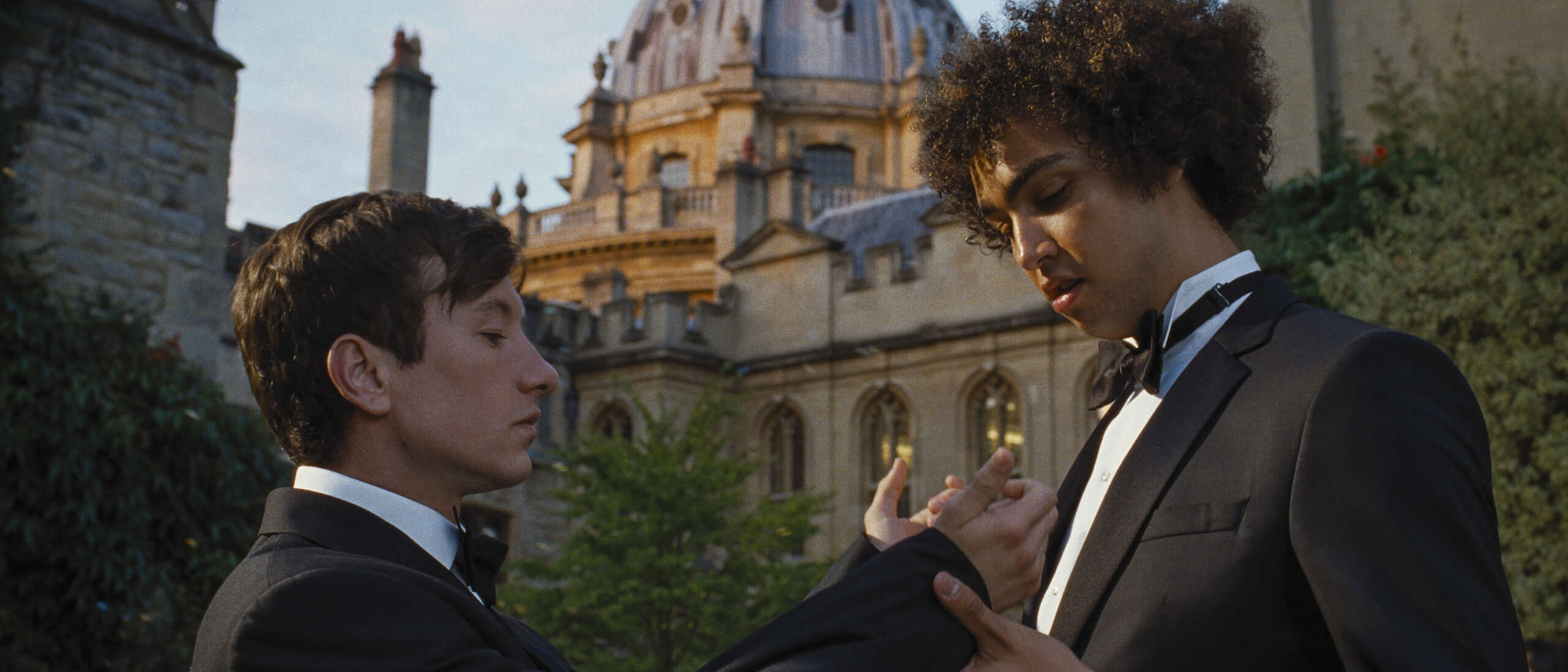 Two men adjust each other's black and white suits outside of an old university building.