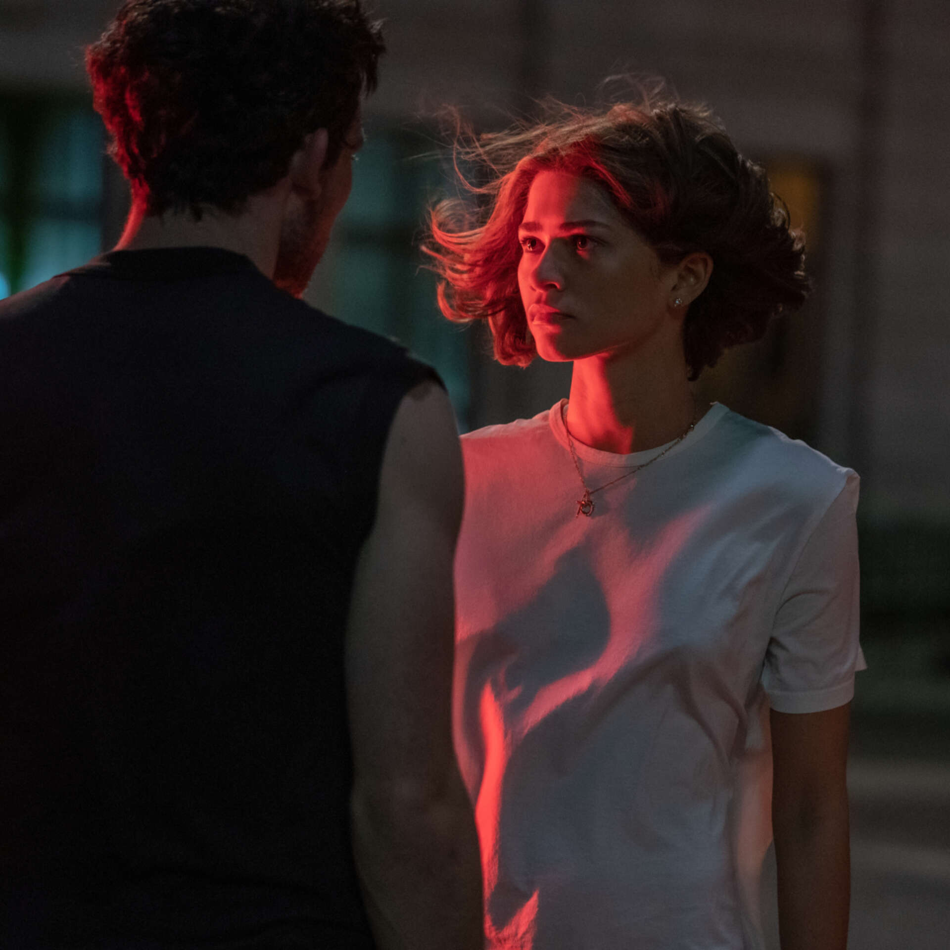 Zendaya and Josh O'Connor star in Challengers. Both are stood facing each other and Josh O'Connor has his back towards the camera. A hot pink light shines on the left side of Zendaya.