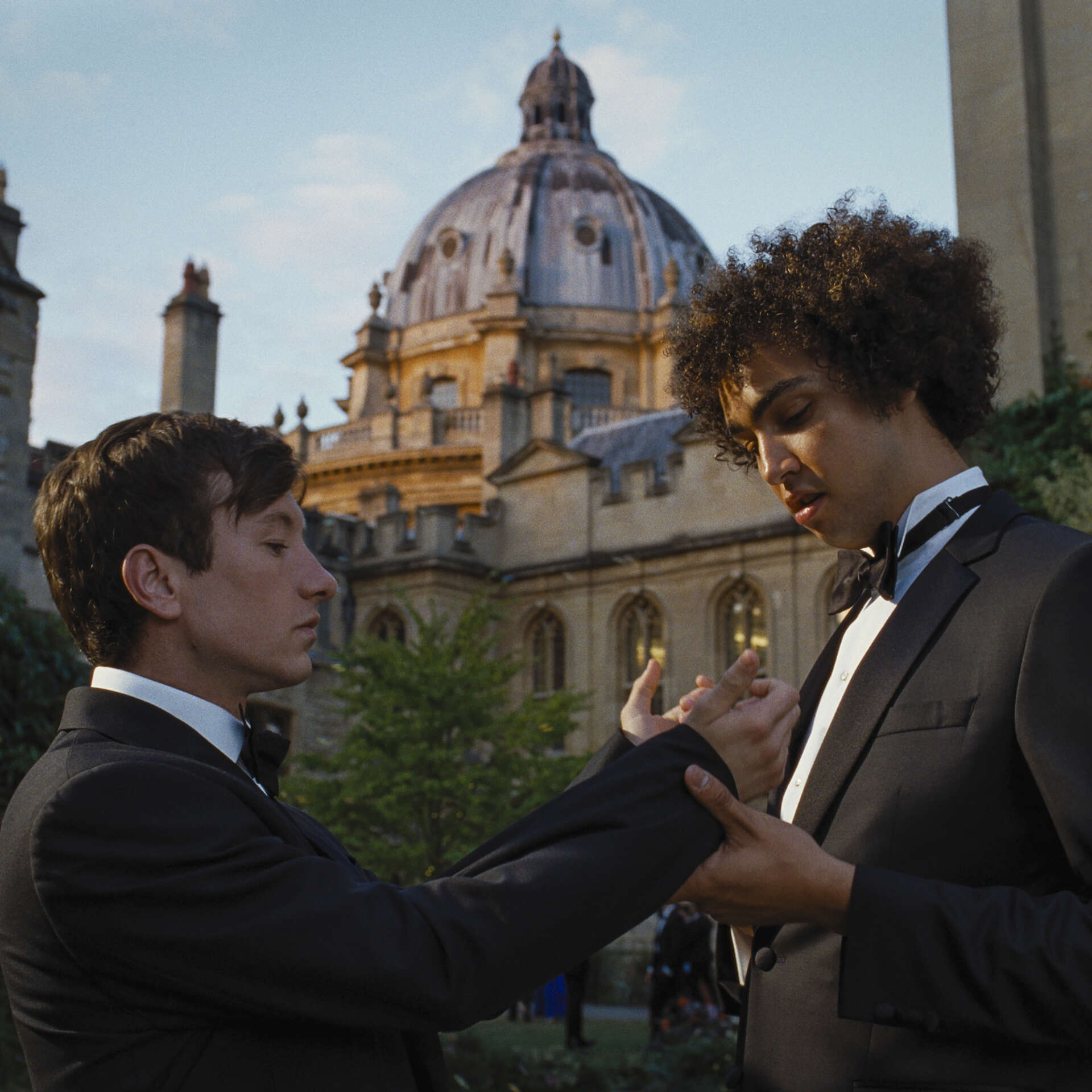 Two men adjust each other's black and white suits outside of an old university building.