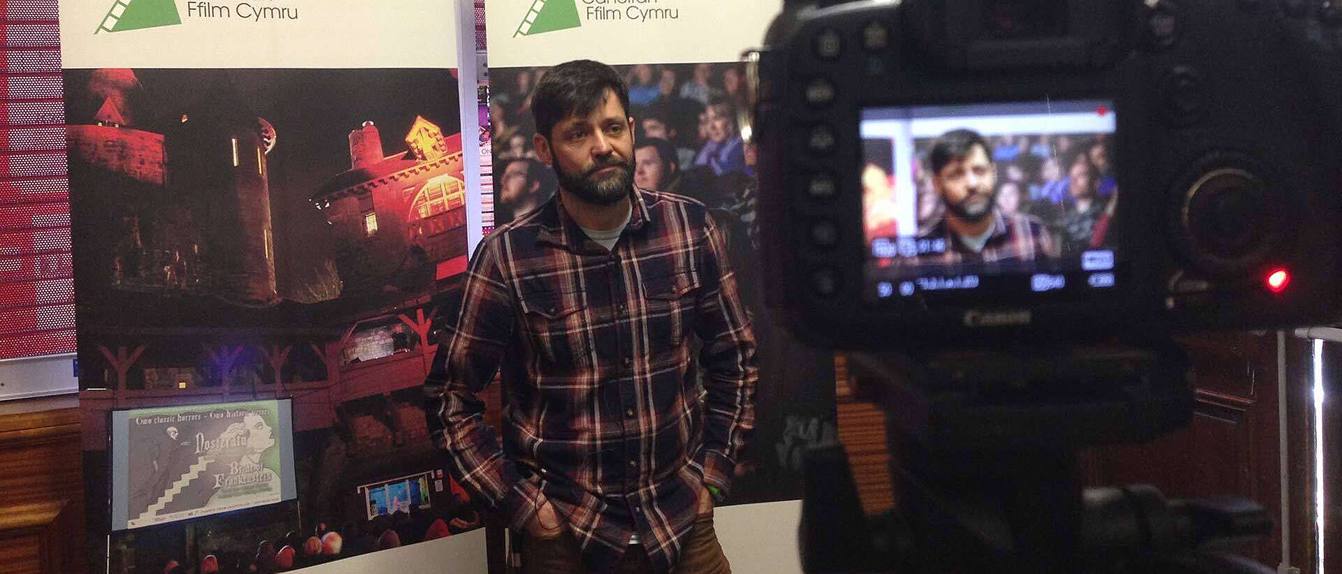A man stands with his hands in his pockets in front of Film Hub Wales posters and poses for a camera.