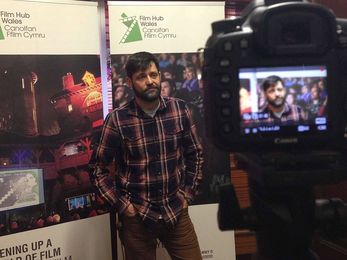 A man stands with his hands in his pockets in front of Film Hub Wales posters and poses for a camera.