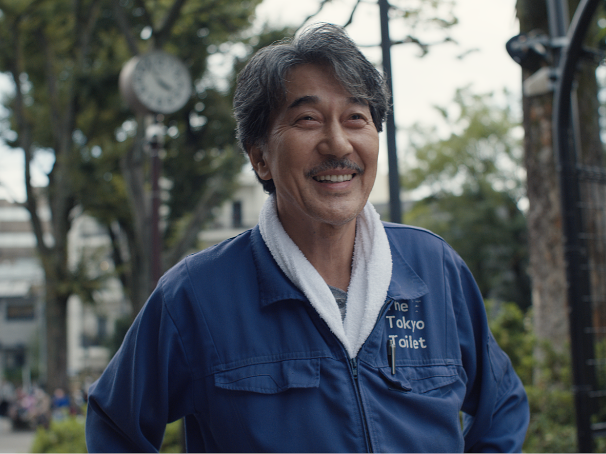 Kôji Yakusho smiles whilst wearing a blue boiler suit in the film Perfect Days.