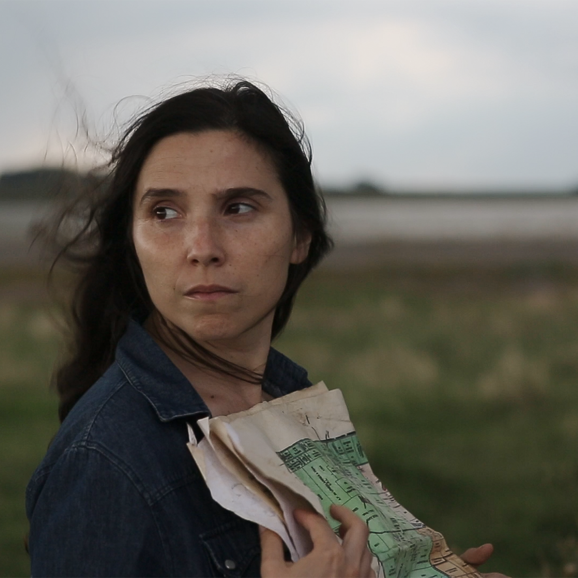 Woman with long dark brown straight hair looks over her right shoulder. She stands in the middle of a green field with the sea in the background and holds a pile of used papers to her chest. Still image from the film Trenque Lauquen I & II.